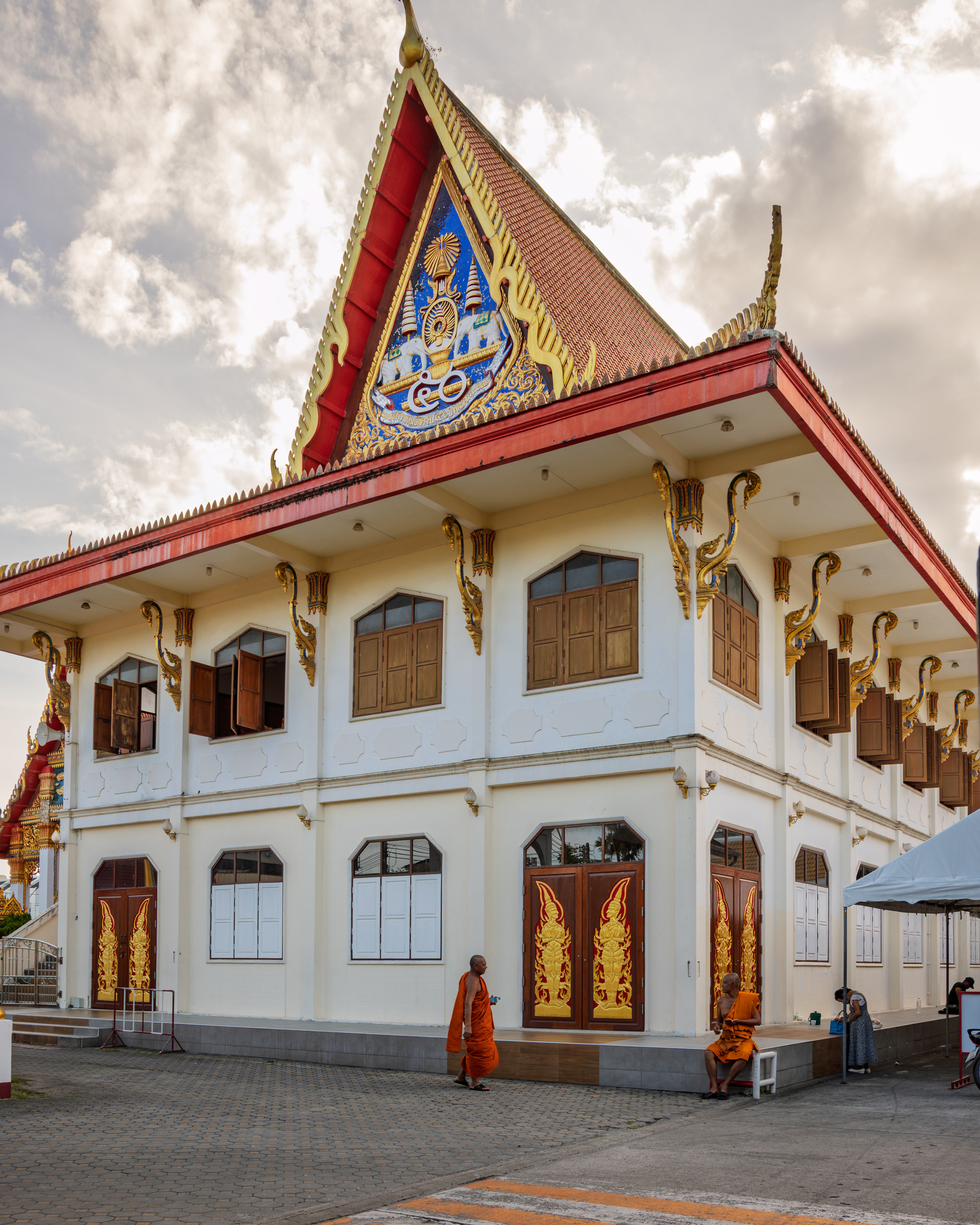 Buddhist temple hall with monks walking past golden-panelled doors and mosaic gable, Thailand — architectural photography
