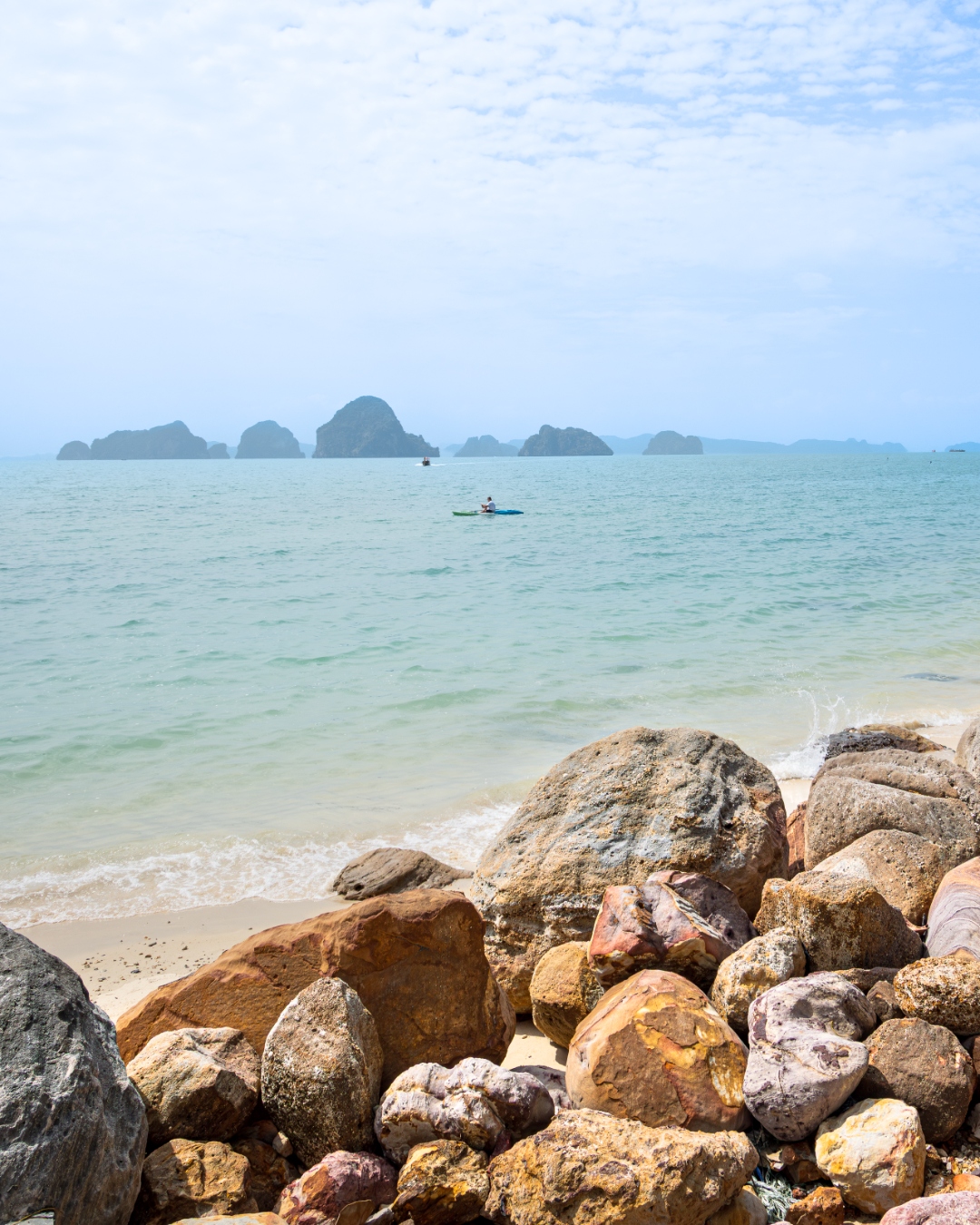 Rocky shoreline and sandy beach with distant karst islands and a lone kayaker, southern Thailand — coastal landscape photography