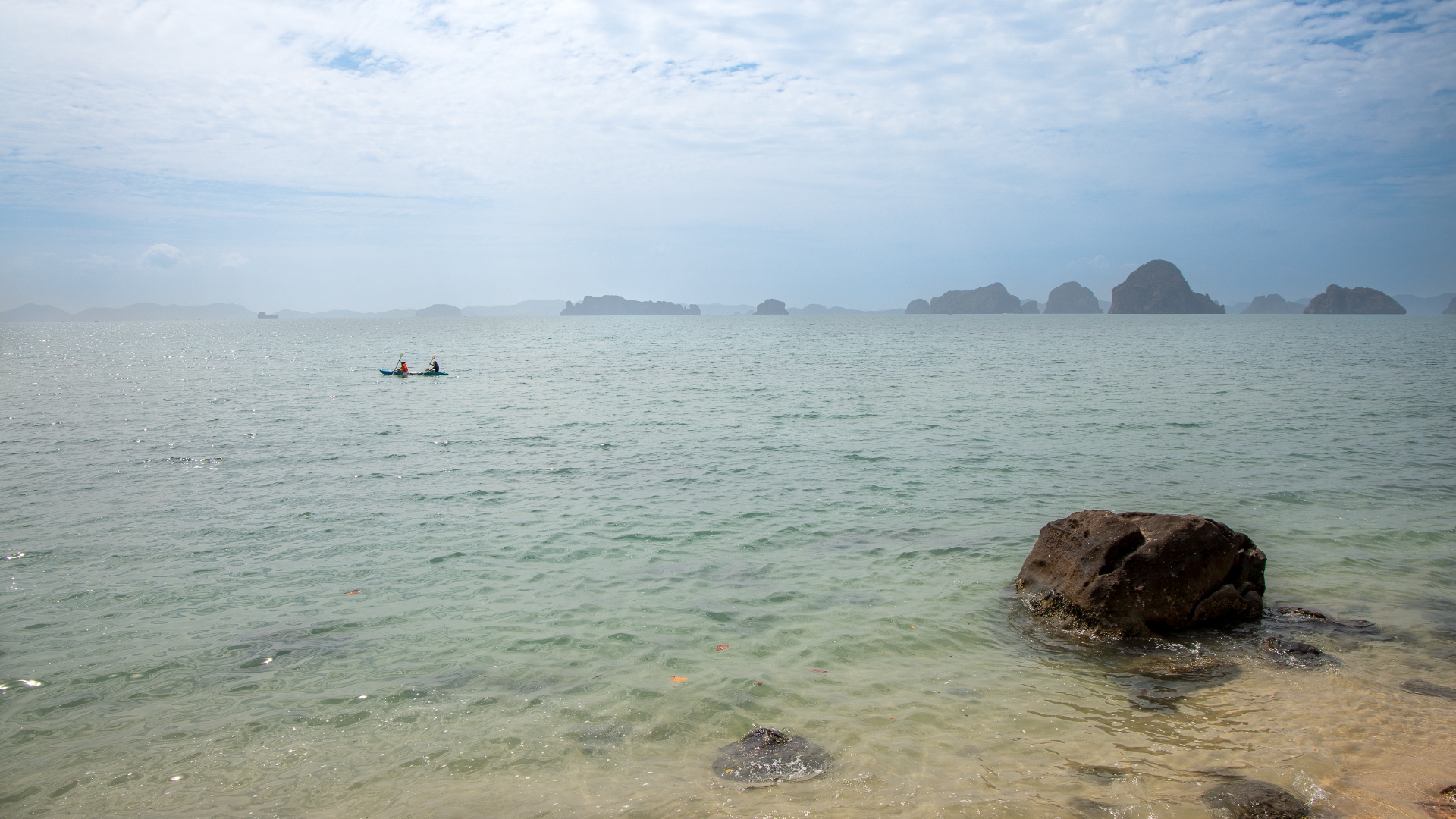 Clear shallow water with a boulder and kayakers paddling towards misty karst islands on the horizon — seascape photography