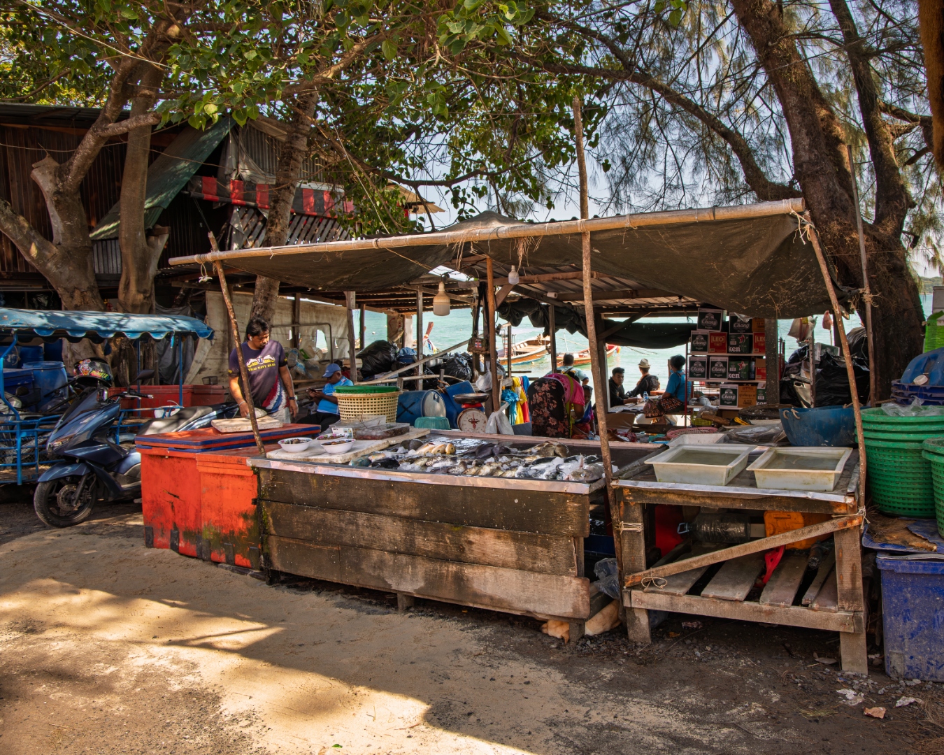 Open-air seaside fish market with vendors selling fresh catch under shade, longtail boats in the harbour — travel photography