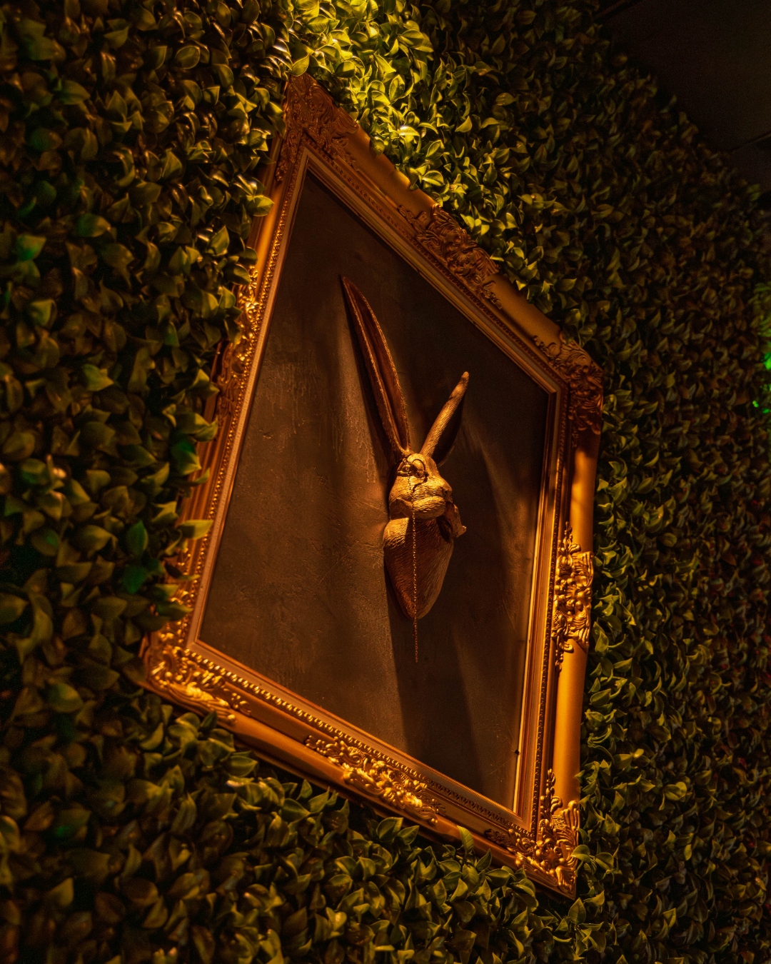 Golden rabbit-head sculpture in ornate gilt frame mounted on a living wall of green leaves — interior photography by Samuel John Ford