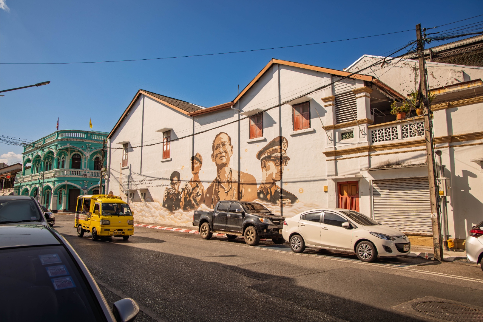Large mural portrait on a white building wall beside a green Sino-Portuguese building, yellow tuk-tuk on the road, Phuket — street photography