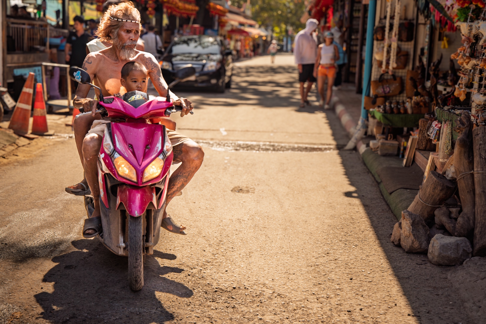 Man and child riding a pink motorbike through golden-hour market street, Thailand — travel photography by Samuel John Ford