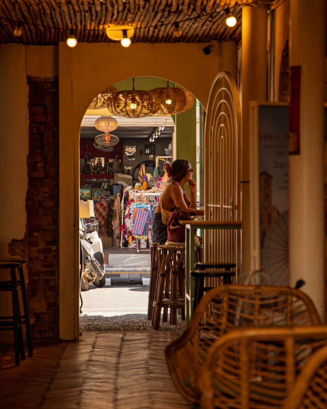 Woman at a rattan bar gazing through an archway into a sunlit street — lifestyle photography, Southeast Asia
